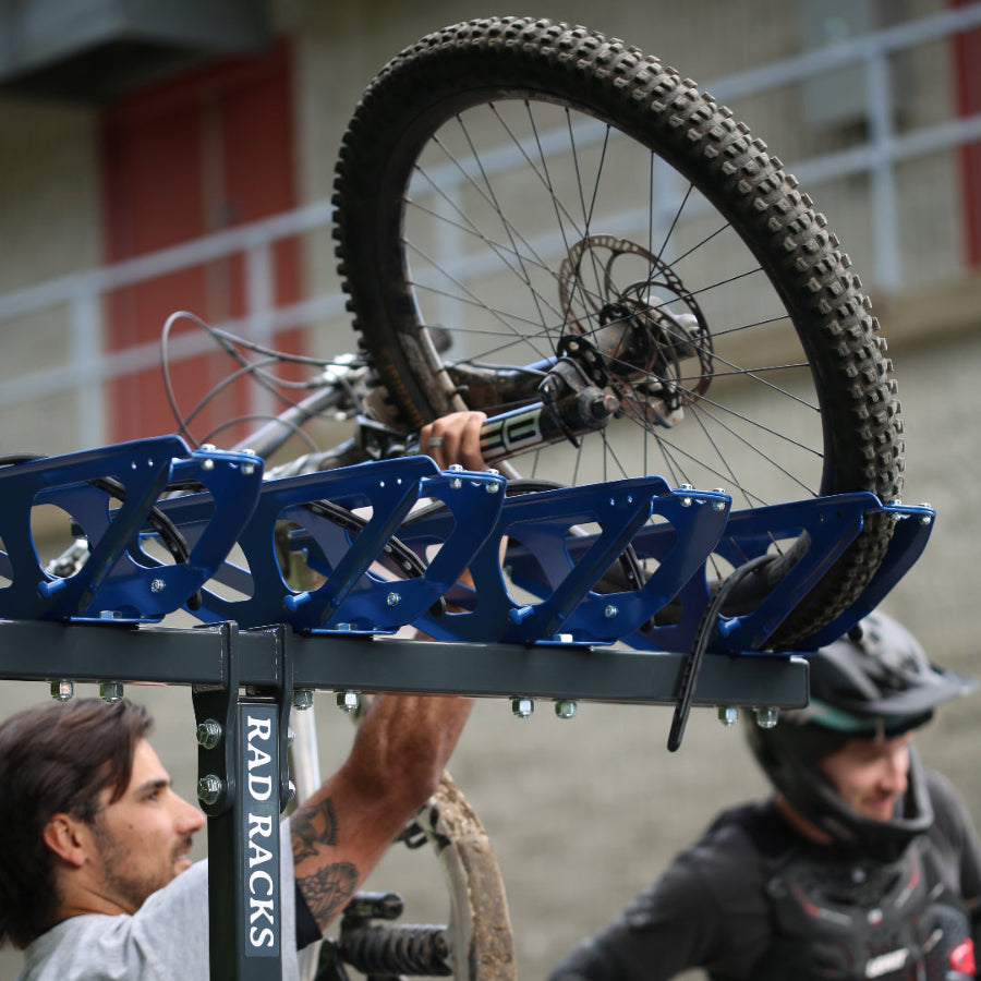 Man loading a mountain bike onto a blue RAD RACKS bike rack mounted on a vehicle, while another person in protective gear stands nearby.