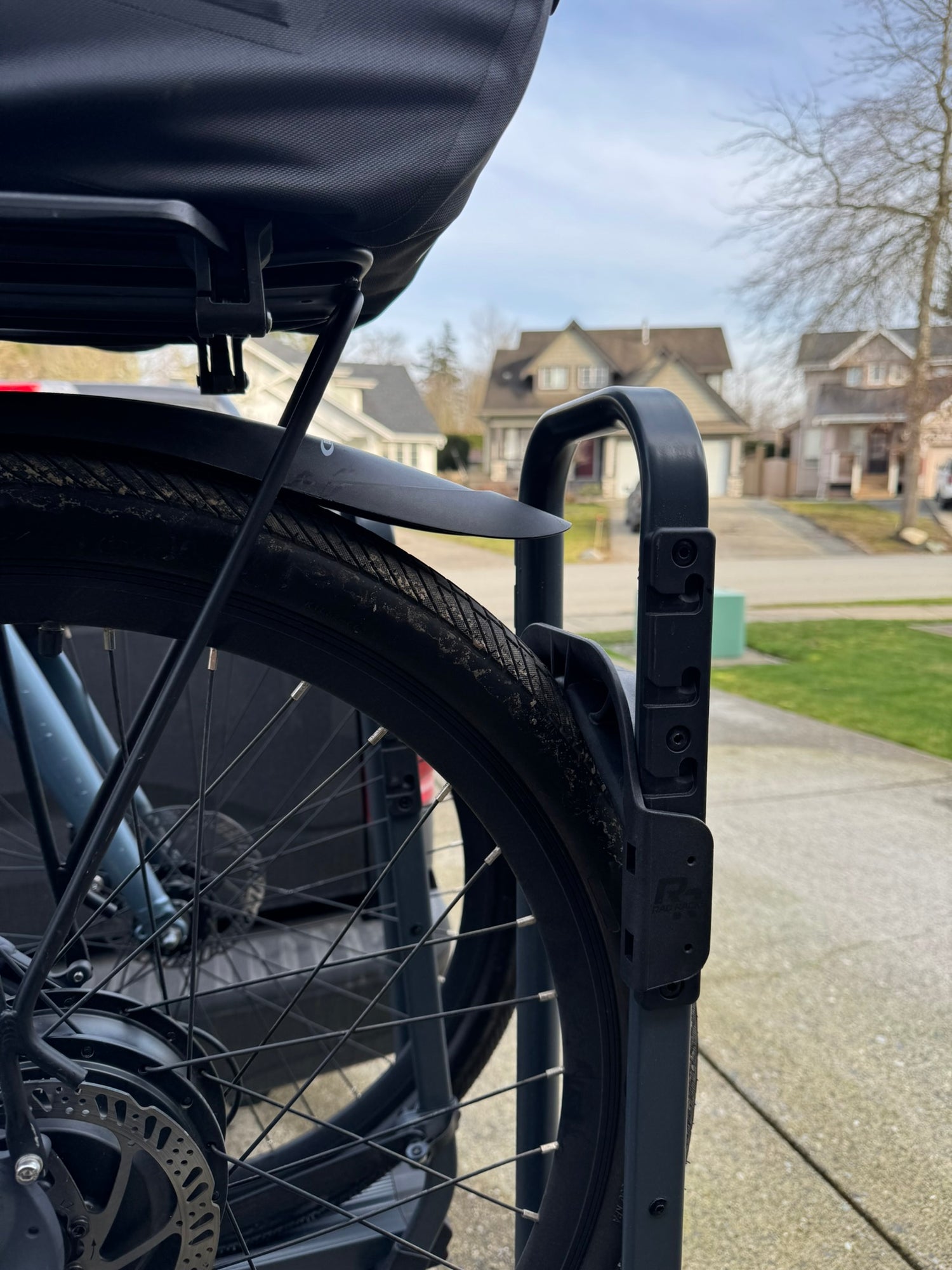 Close-up of a bicycle wheel with a black tire and rim, mounted on a bike trailer, with a residential area in the background. Bike rack clears fenders, tray bike rack
