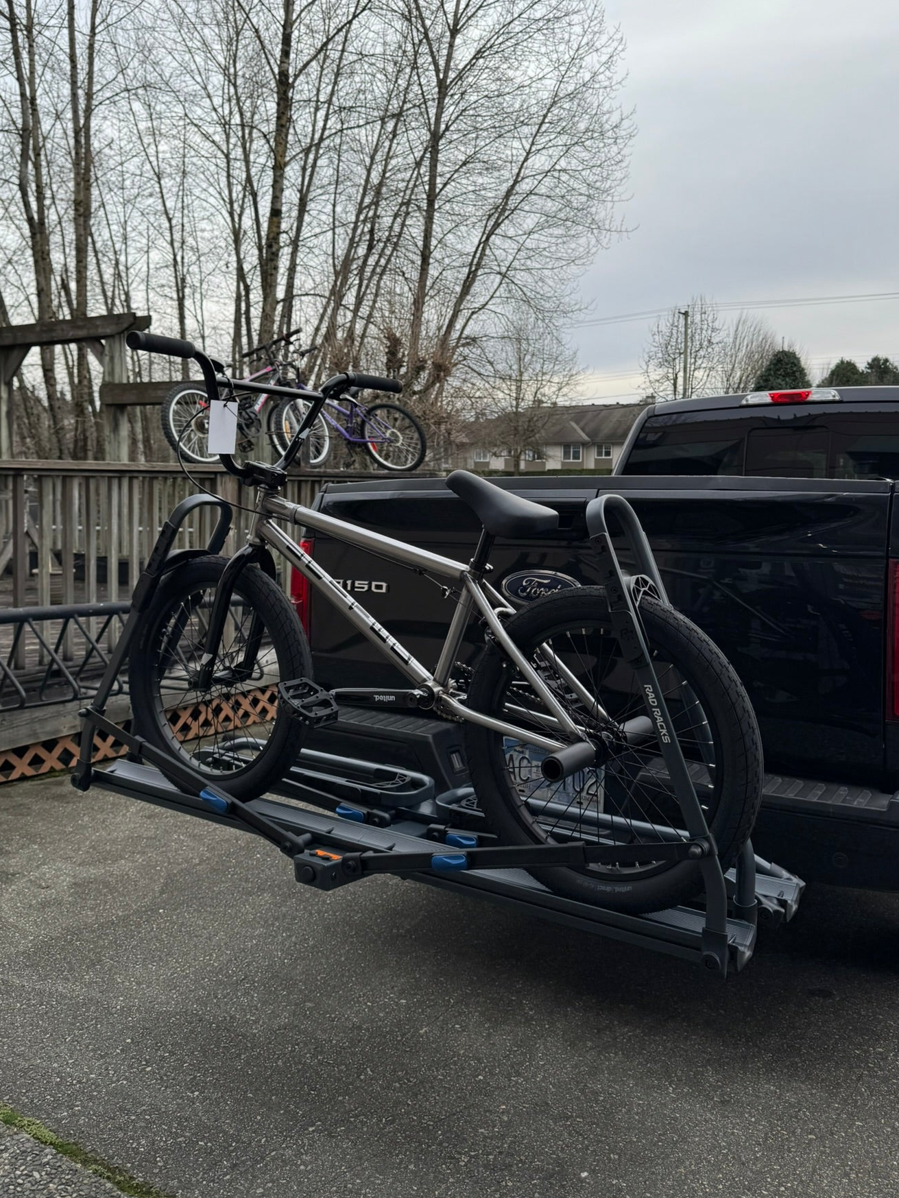 BMX bicycle on a bike rack attached to the back of a pickup truck with leafless trees and a cloudy sky in the background.