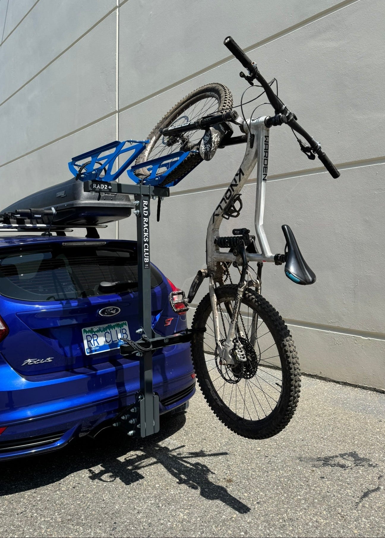 Blue car with a bike mounted on a rack against a gray wall.
