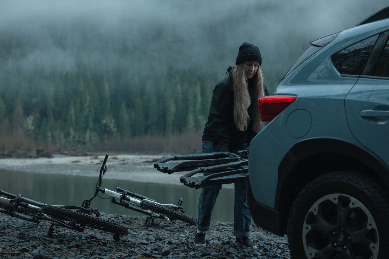 Person with bicycles near a car by a lake with mountains in the background